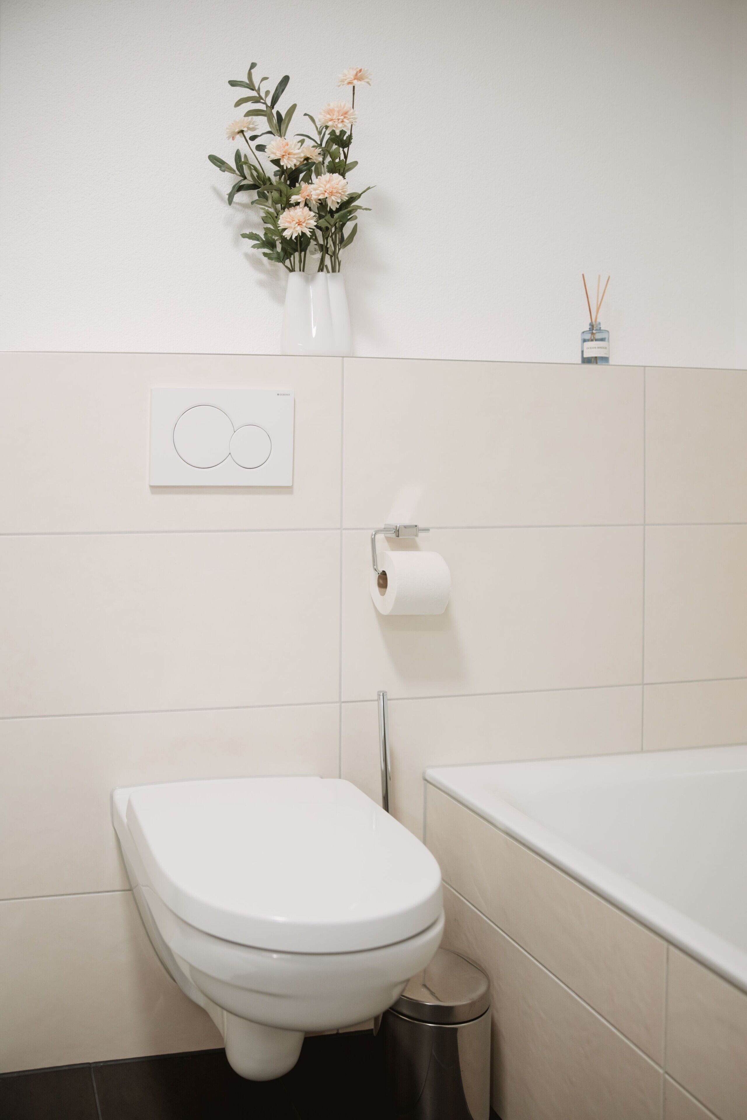 Une salle de bain comprenant des toilettes, un bidet, une poubelle, un porte-rouleau de papier toilette, un arrangement floral fixé au mur et un petit flacon de diffuseur de parfum à bâtonnets.