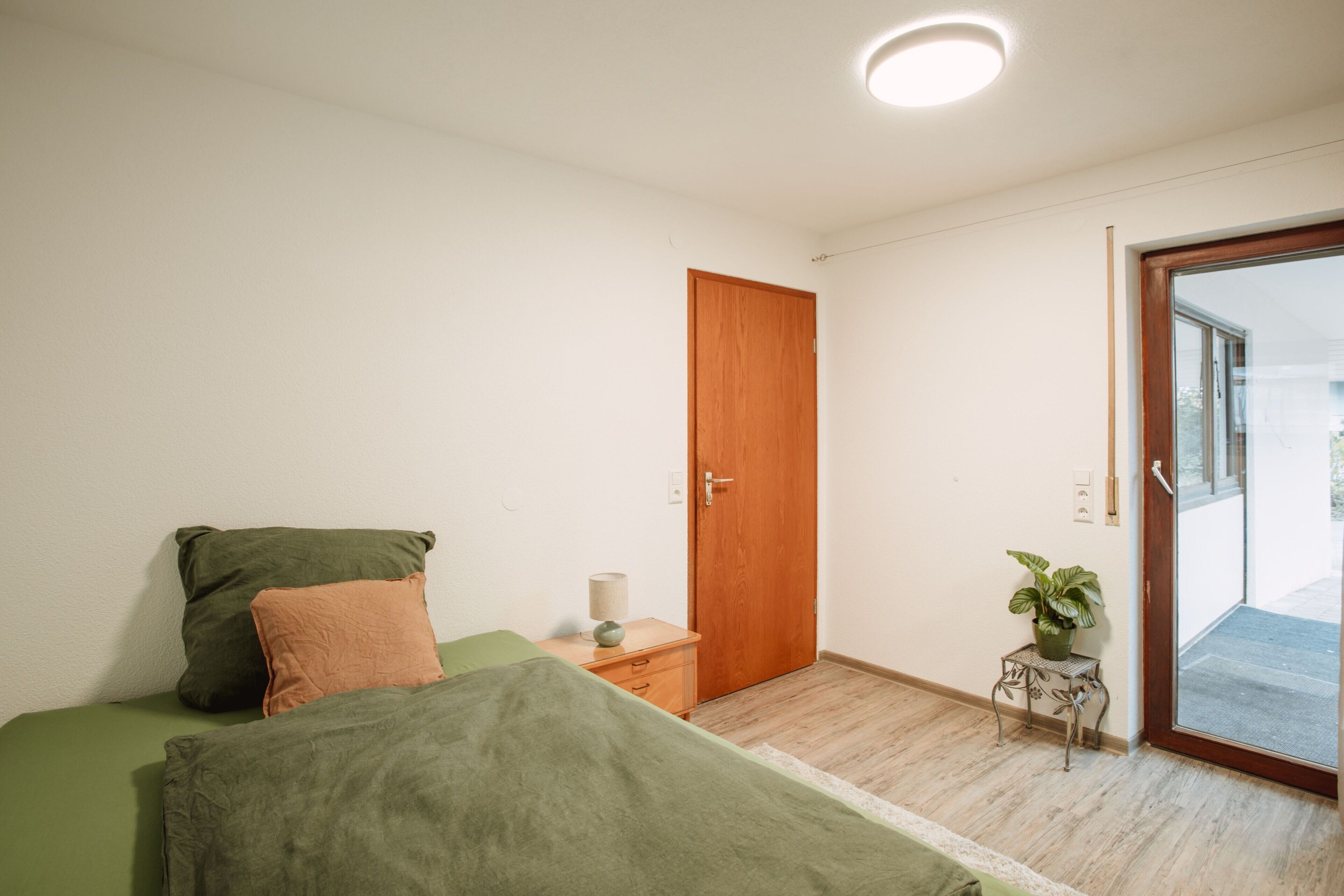 Un dormitorio con una cama, una puerta de madera, una mesita con una planta y vistas a través de una puerta de cristal.