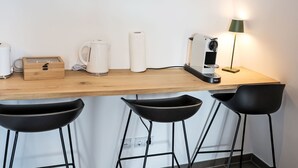 Una kitchenette con cafetera, hervidor y una caja de madera. Hay tres taburetes negros y una lámpara de pie verde.