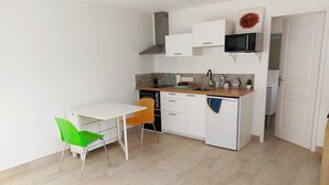 Cocina compacta con armarios blancos, microondas y fregadero. Hay una pequeña mesa de comedor con dos sillas, una verde y otra naranja.