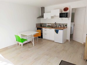 Eine kompakte Küche mit weißen Schränken, einer Mikrowelle und einem Spülbecken. Es gibt einen kleinen Esstisch mit zwei Stühlen, einem grünen und einem orangefarbenen.