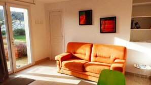 Una sala moderna con un sofá naranja grande, un sillón verde y una mesita auxiliar pequeña. Hay dos cuadros enmarcados en la pared y una puerta que da a un jardín.