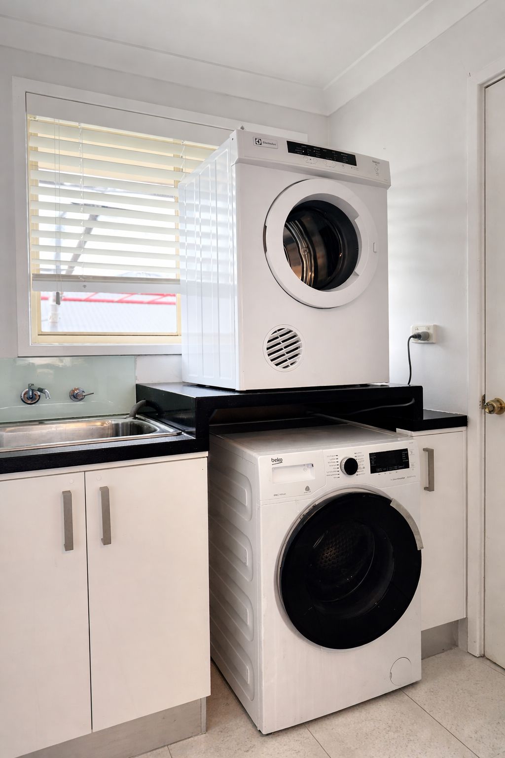 Une petite buanderie équipée d’un lave-linge et d’un sèche-linge à chargement frontal, d’un évier et d’une fenêtre avec des stores.