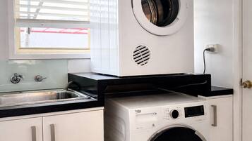Une petite buanderie équipée d’un lave-linge et d’un sèche-linge à chargement frontal, d’un évier et d’une fenêtre avec des stores.