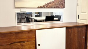Ein Holzschrank mit weißer Tür, darüber ein Gemälde mit Strandmotiv und darunter die Spiegelung eines Essbereichs.