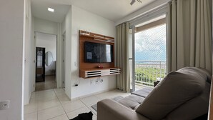 A living room with a flat-screen TV mounted on a wooden console, a sofa, and a balcony with a view. - Apto. Novo Perto da Praia da Ponta Negra (Manaus)