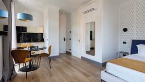 Quarto de hotel moderno com cama, área de jantar com cadeiras e mesa, cozinha com eletrodomésticos e banheiro com espelho.