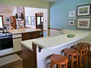 A kitchen with a large island, wooden stools, a sink, and a stove. - Gray House at Pawleys - 3 bedroom - Pool (Pawleys Island)