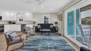 A modern kitchen with white cabinets, a ceiling fan, and a dining area with a black sofa and a blue rug. - Haney's By The C - Unit C of Triplex (Pawleys Island)