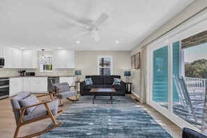 A modern kitchen with white cabinets, a ceiling fan, and a dining area with a black sofa and a blue rug. - Haney's By The C - Unit C of Triplex (Pawleys Island)