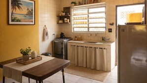Eine kleine Küche mit Gasherd, Spüle und Kühlschrank. Ein Fenster mit Jalousien, eine kleine Ablage mit Dekoration und ein Bild an der Wand.