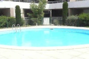 Une piscine avec un escalier en métal, entourée de verdure et bordée d’un bâtiment doté de plusieurs balcons.