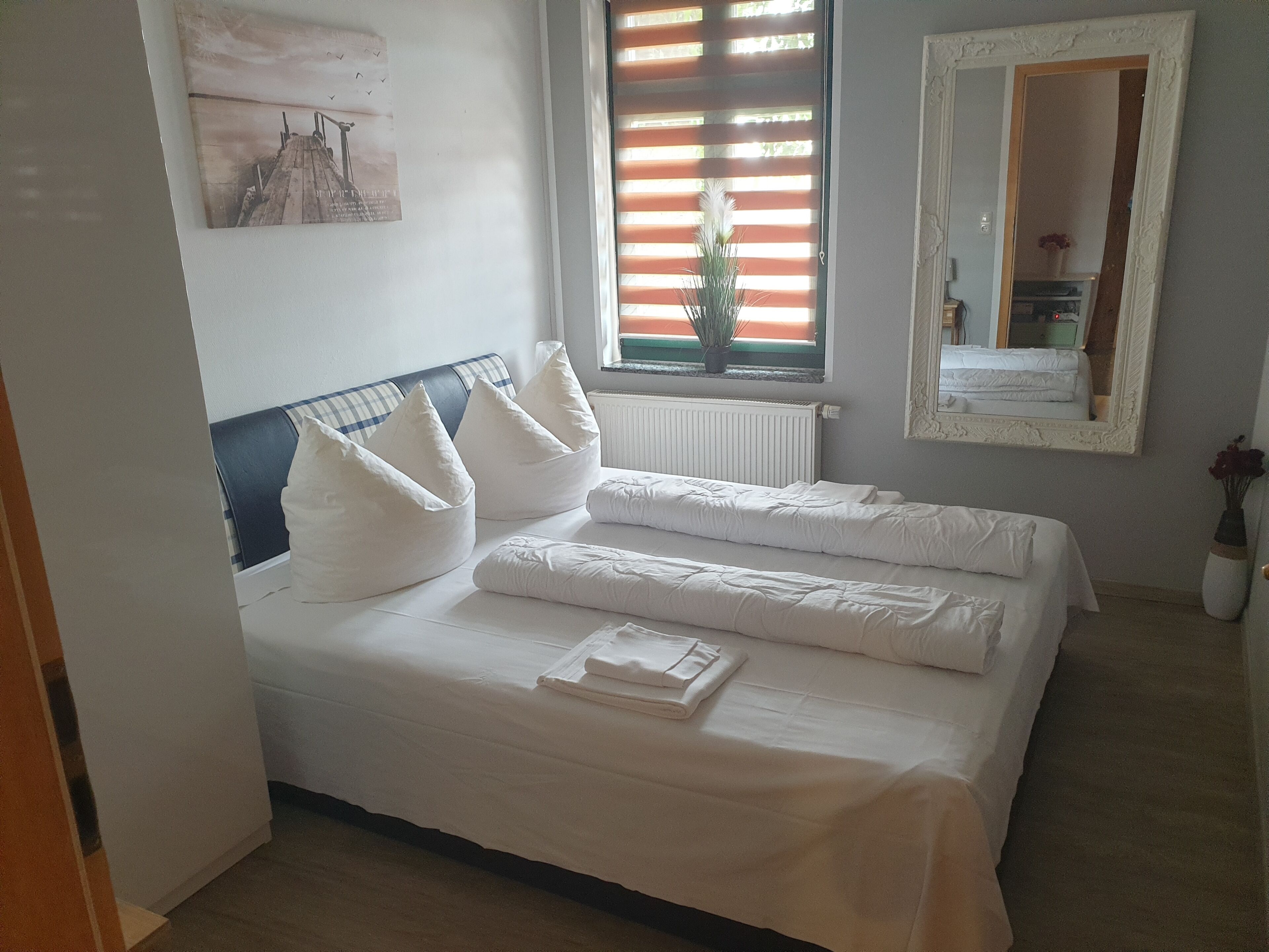 Een opgemaakt bed met witte lakens en kussens, een raam met zonwering, een radiator, een spiegel en een schilderij aan de muur.