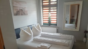 Una cama bien hecha con sábanas y almohadas blancas, una ventana con persianas, un radiador, un espejo y un cuadro en la pared.