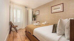A bedroom with a wooden floor, a bed with white bedding, a wooden chair, a nightstand with a vase, and a framed picture on the wall. - A janela de Alfama (Lisbon)