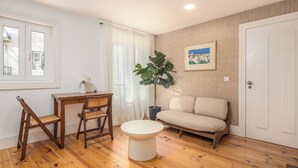 A room with a wooden floor, a round white table, two wooden chairs, a beige sofa, a potted plant, a framed picture on the wall, and a window with white curtains. - A janela de Alfama (Lisbon)