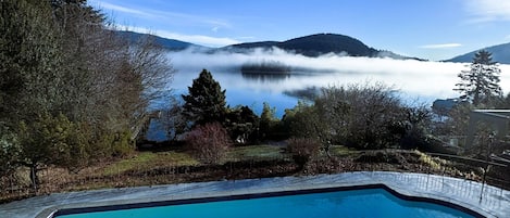 A swimming pool with clear blue water, surrounded by a landscaped garden with trees and a view of a lake and mountains in the background.