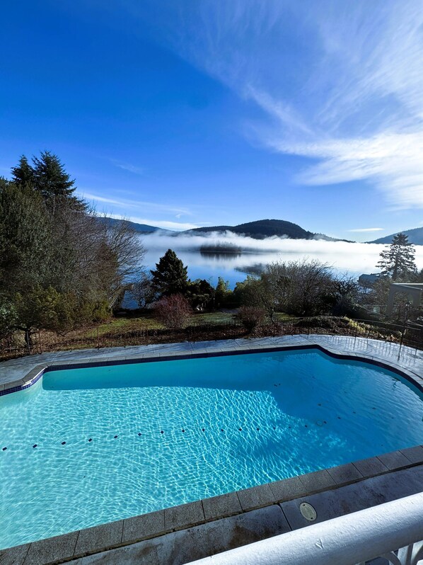 Uma piscina com água azul cristalina, rodeada por um jardim ajardinado com árvores e vista para um lago e montanhas ao fundo.