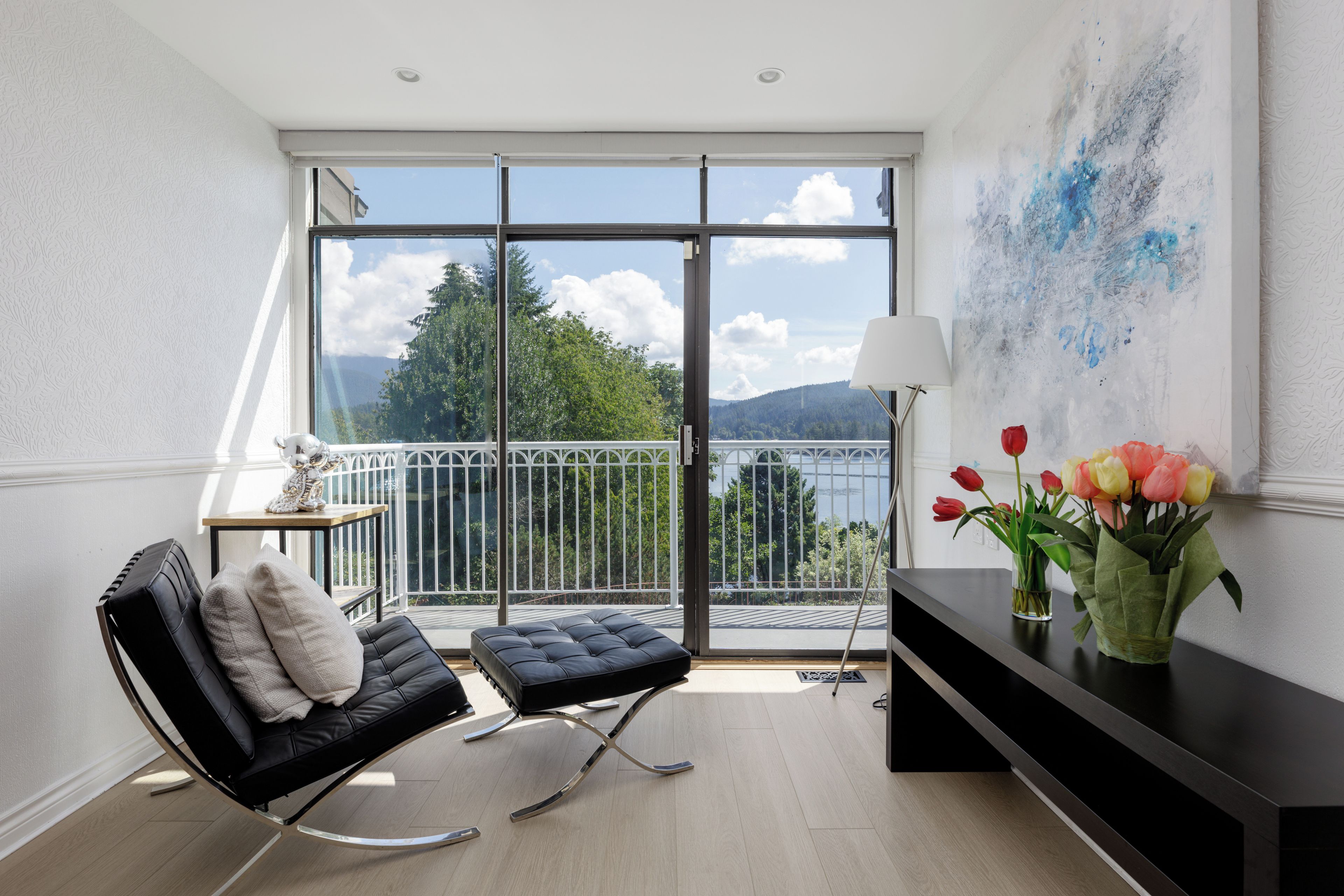 Een moderne kamer met een zwarte lederen fauteuil, een klein tafeltje met een lamp en uitzicht op een meer door grote ramen.