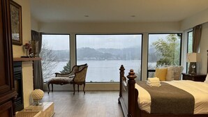 Une chambre spacieuse avec un grand lit, un fauteuil et une vue sur un lac, grâce à de grandes fenêtres.