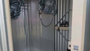 A compact, enclosed space with a metal corrugated wall, a white toilet, and a shelf with items. - Vintage Bedford Bus Farm Glamping Under the Stars (Silverwood)