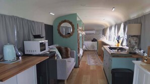 A compact kitchen with a microwave, kettle, and sink. There is a small dining area with a table and chairs. The room has a light blue wall and a wooden floor. - Vintage Bedford Bus Farm Glamping Under the Stars (Silverwood)