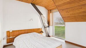 Een slaapkamer met een houten schuine balkenlaag, een bed met een witte matras en een houten hoofdbord, een nachtkastje met een lamp, en een raam met zonwering.