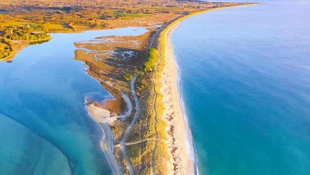Luchtbeeld van een kustgebied met een strand, helderblauw water en een combinatie van natuurlijke en bebouwde gebieden.