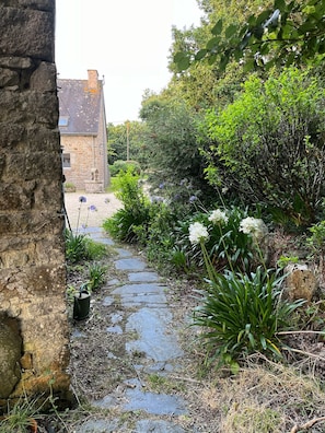 Un chemin de pierres traverse un jardin où poussent différentes espèces de plantes, et un bâtiment en pierre se dresse à l’arrière-plan.