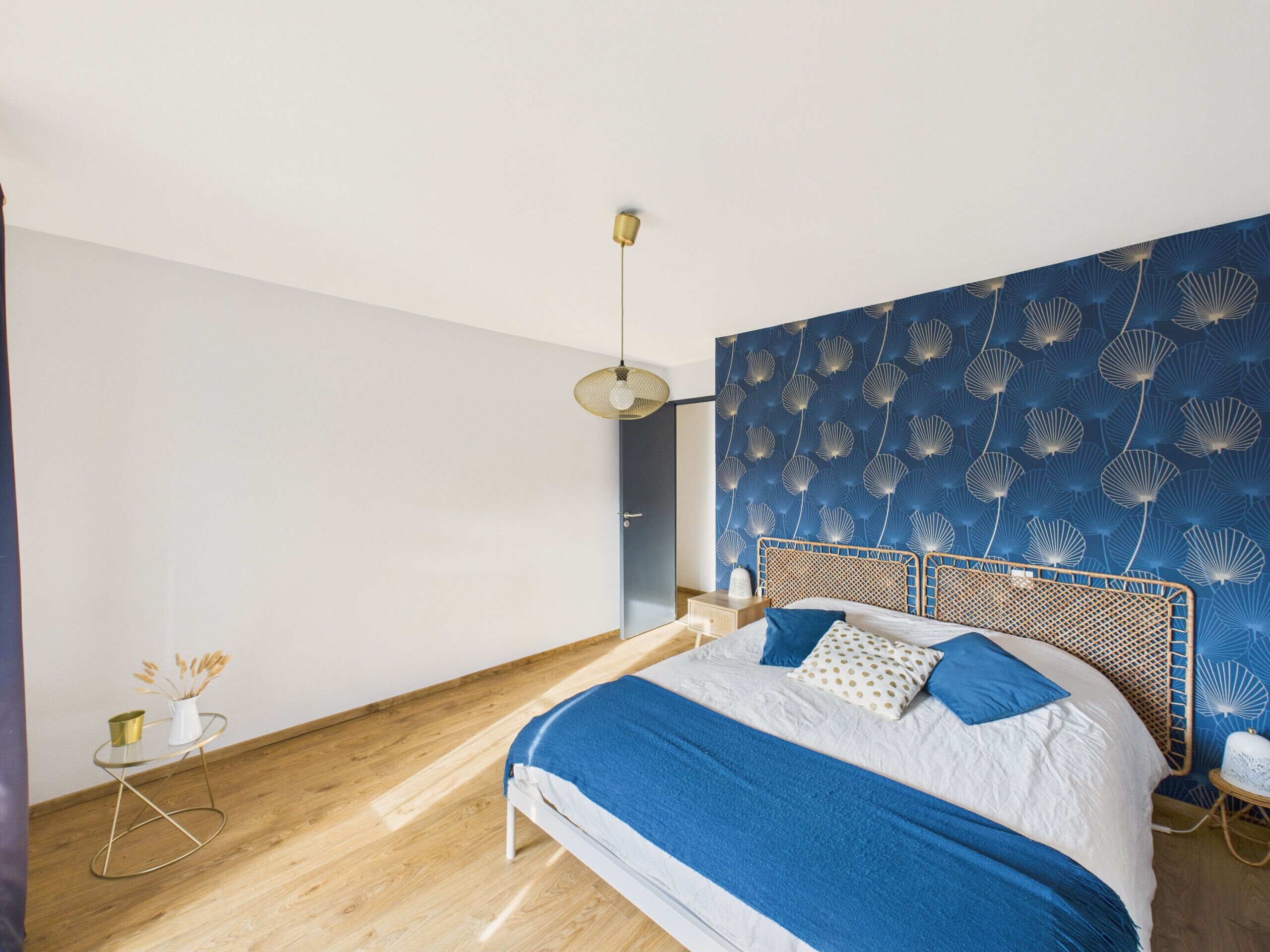 Une chambre à coucher avec un lit, une petite table sur laquelle est posé un vase, une lampe suspendue et un mur orné d’un motif de coquillages.