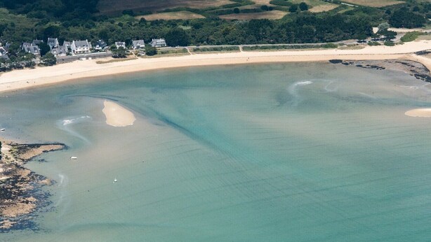 An aerial view of a coastal area with a sandy beach, clear water, and a cluster of houses in the background.