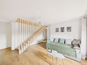 A modern living room with a wooden staircase, a sofa with cushions, a small table, and wall art. - City center – Large house for 10 with garden (Ploemel)