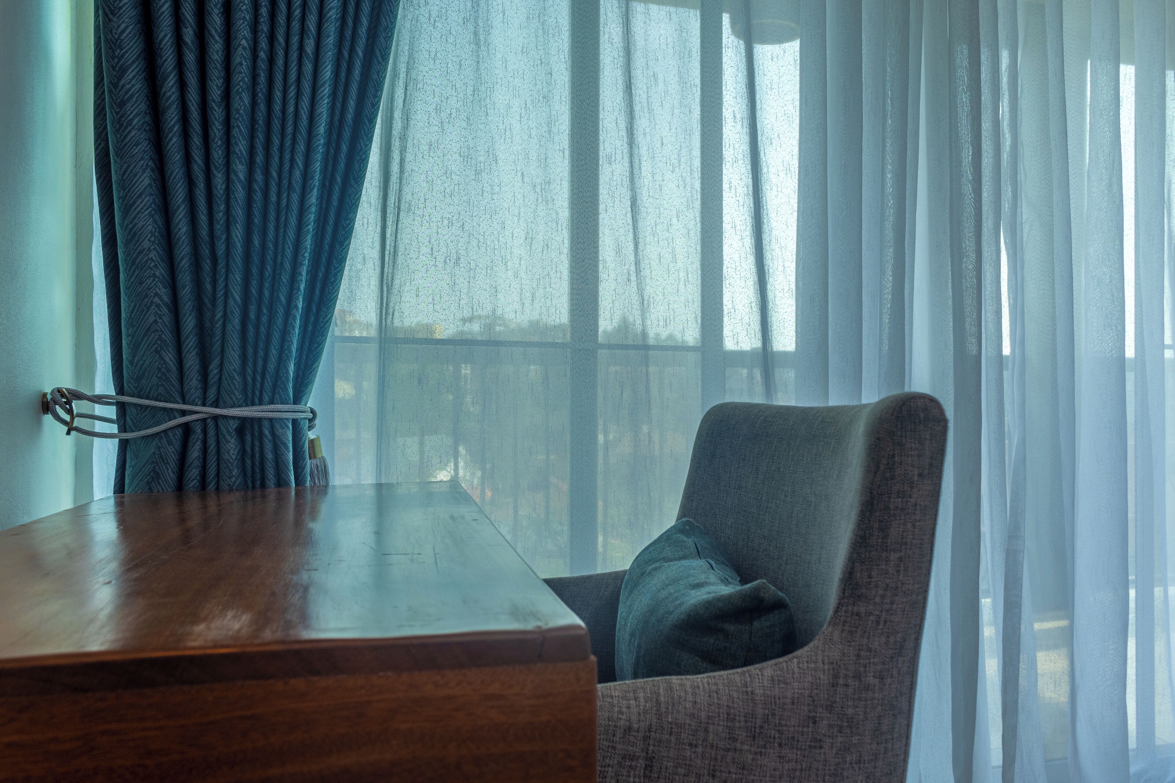 A room with a wooden desk, a grey armchair, and sheer curtains.