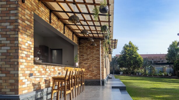 A patio area with a brick wall, wooden chairs, hanging planters, and a view of a green lawn and trees. - VALLEY BLOOM HOTEL KYANJA (Kampala)