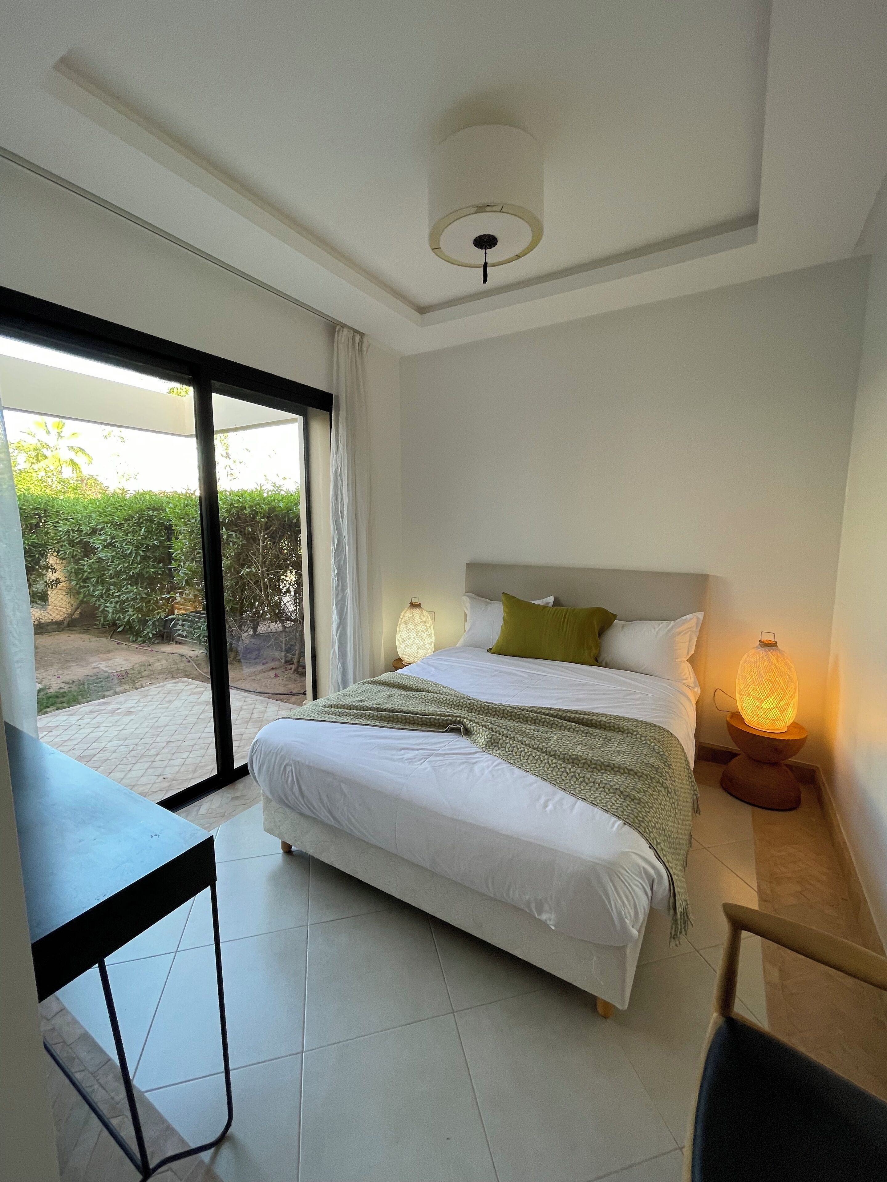 A bedroom with a bed, a desk, a chair, a nightstand with a lamp, and a view of the outdoors through a sliding door.