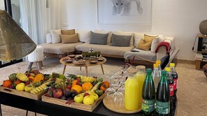 Ein Wohnzimmer mit einer Couch, einem Tisch mit Früchten und Getränken sowie einem gerahmten Bild an der Wand.