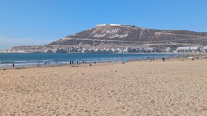 Ein Strand mit einer großen Hügelkette im Hintergrund, bewölktem Himmel und einigen Personen sowie Sonnenschirmen im Sand.
