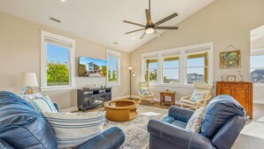 A spacious living room with a ceiling fan, large windows, and a view of a house. - Brand New Semi-Oceanfront Waves Home with Private Pool, Hot Tub, and Elevator (Waves)