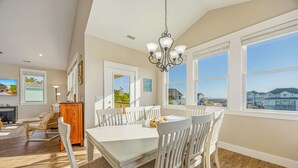 A spacious dining area with a white table and chairs, large windows offering a view of the sea and other buildings, and a chandelier hanging from the ceiling. - Brand New Semi-Oceanfront Waves Home with Private Pool, Hot Tub, and Elevator (Waves)