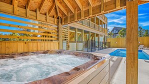 A hot tub with a wooden deck, a pool, and a two-story building with large windows. - Brand New Semi-Oceanfront Waves Home with Private Pool, Hot Tub, and Elevator (Waves)