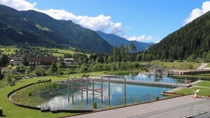 Ein Schwimmbad mit einer umlaufenden Sonnenterrasse, einer kleinen Brücke und Blick auf eine gebirgige Landschaft mit einem Dorf im Hintergrund.
