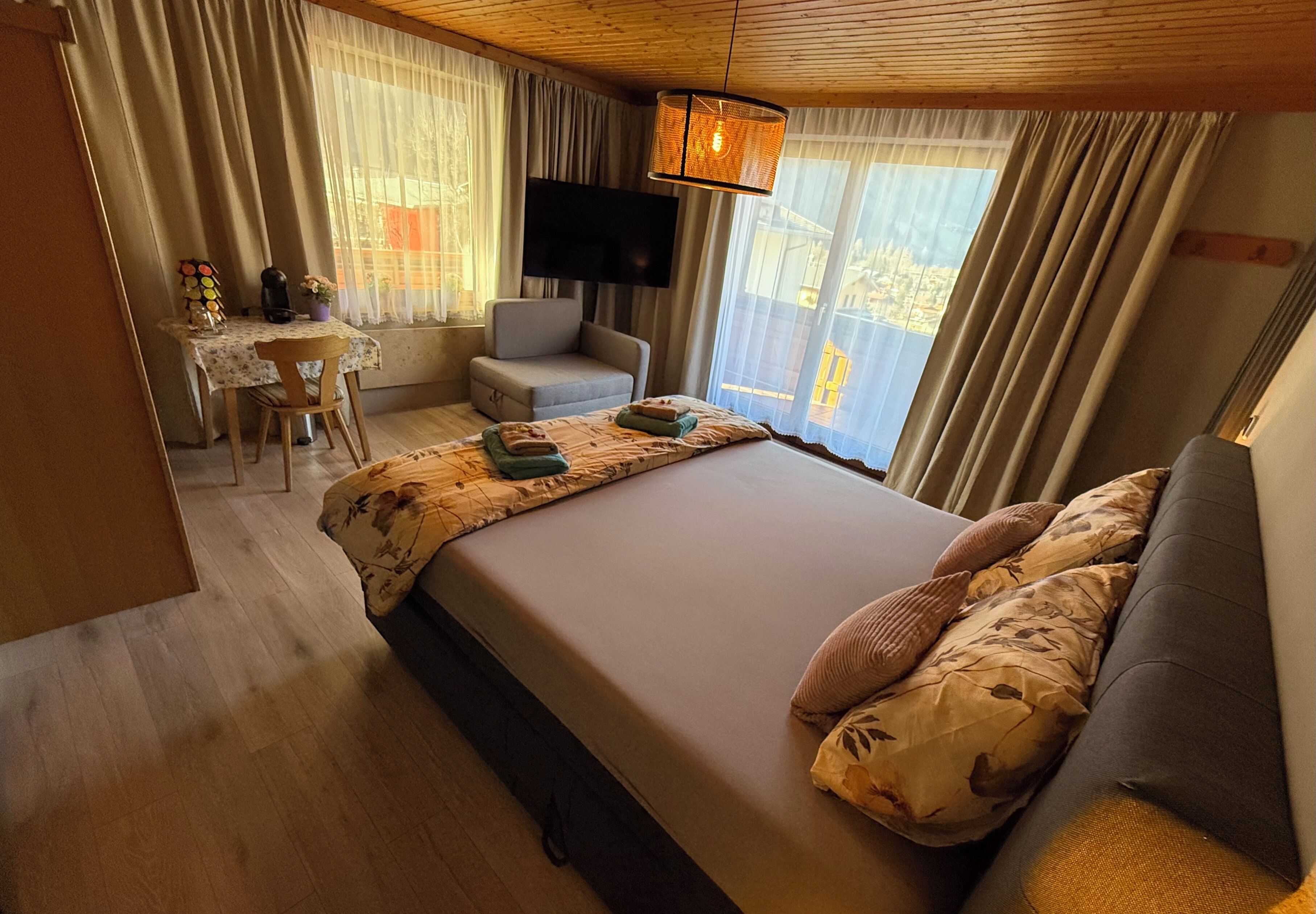 A hotel room with a bed, a chair, a small table, a TV, and a balcony with curtains.