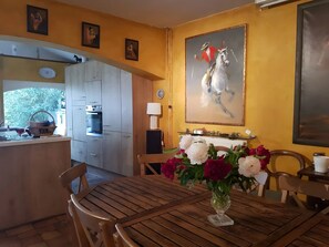 A dining area with a wooden table, chairs, a vase of flowers, a painting, and a kitchen with cabinets and appliances.