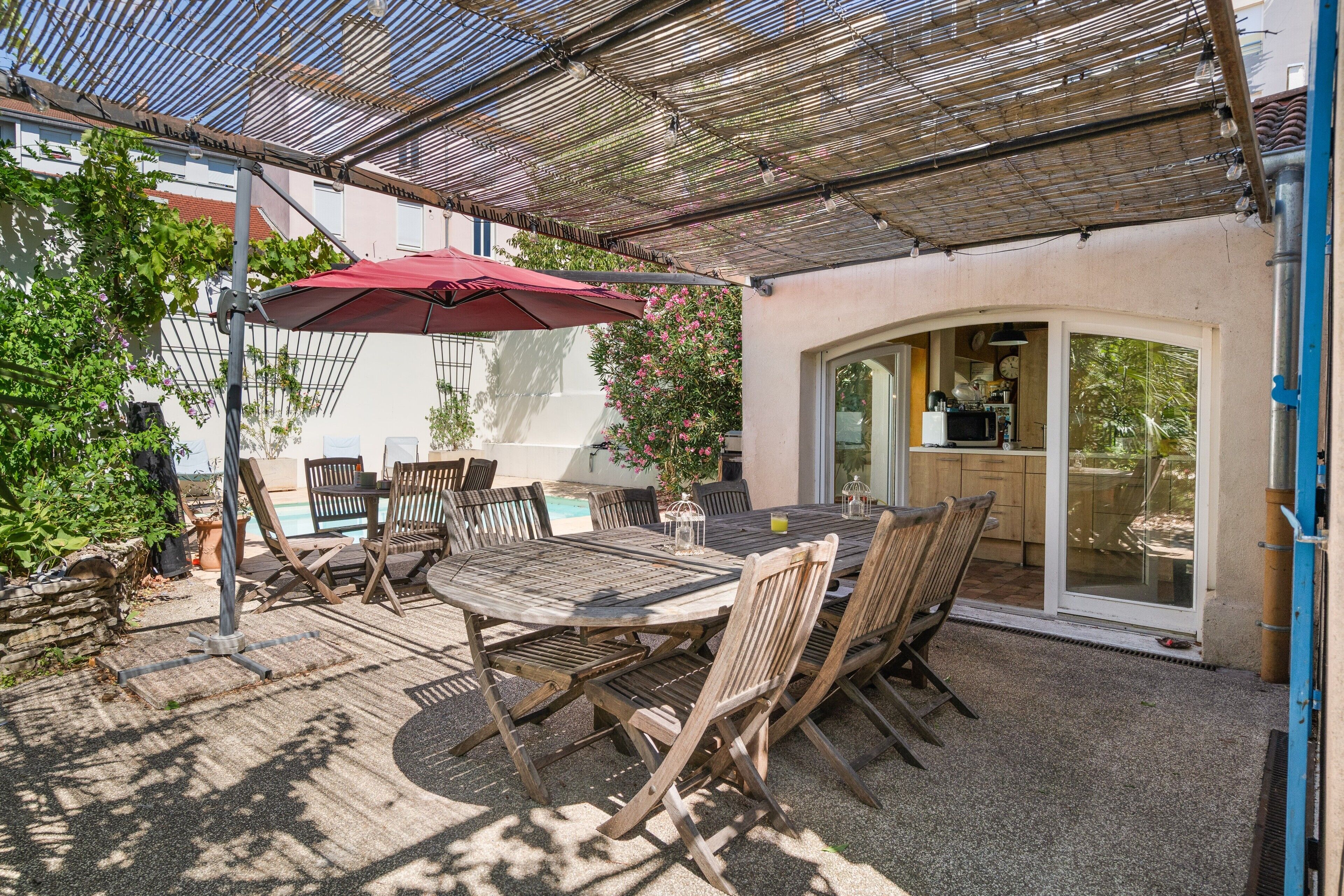Een terras buiten met een tafel en stoelen, een rode parasol, en uitzicht op een keuken door een glazen deur.