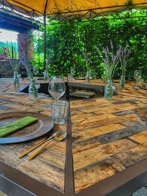 Uma área de refeições ao ar livre com mesas de madeira, copos de vidro transparente e lavanda em vasos.