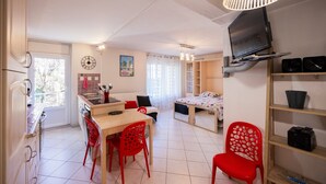 Una camera compatta con angolo cottura, zona pranzo, un letto, una TV e uno scaffale con dispositivi elettronici.