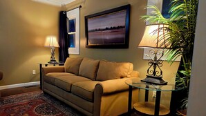 A living room with a beige sofa, a glass coffee table, a patterned rug, a framed landscape painting, a lamp, and a potted plant.