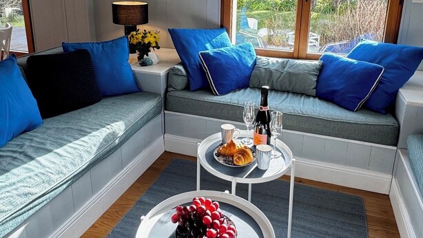 Een moderne woonkamer met een grijze bank, blauwe kussens, een klein rond tafeltje met een fruitschaal, en een raam met uitzicht op groen.