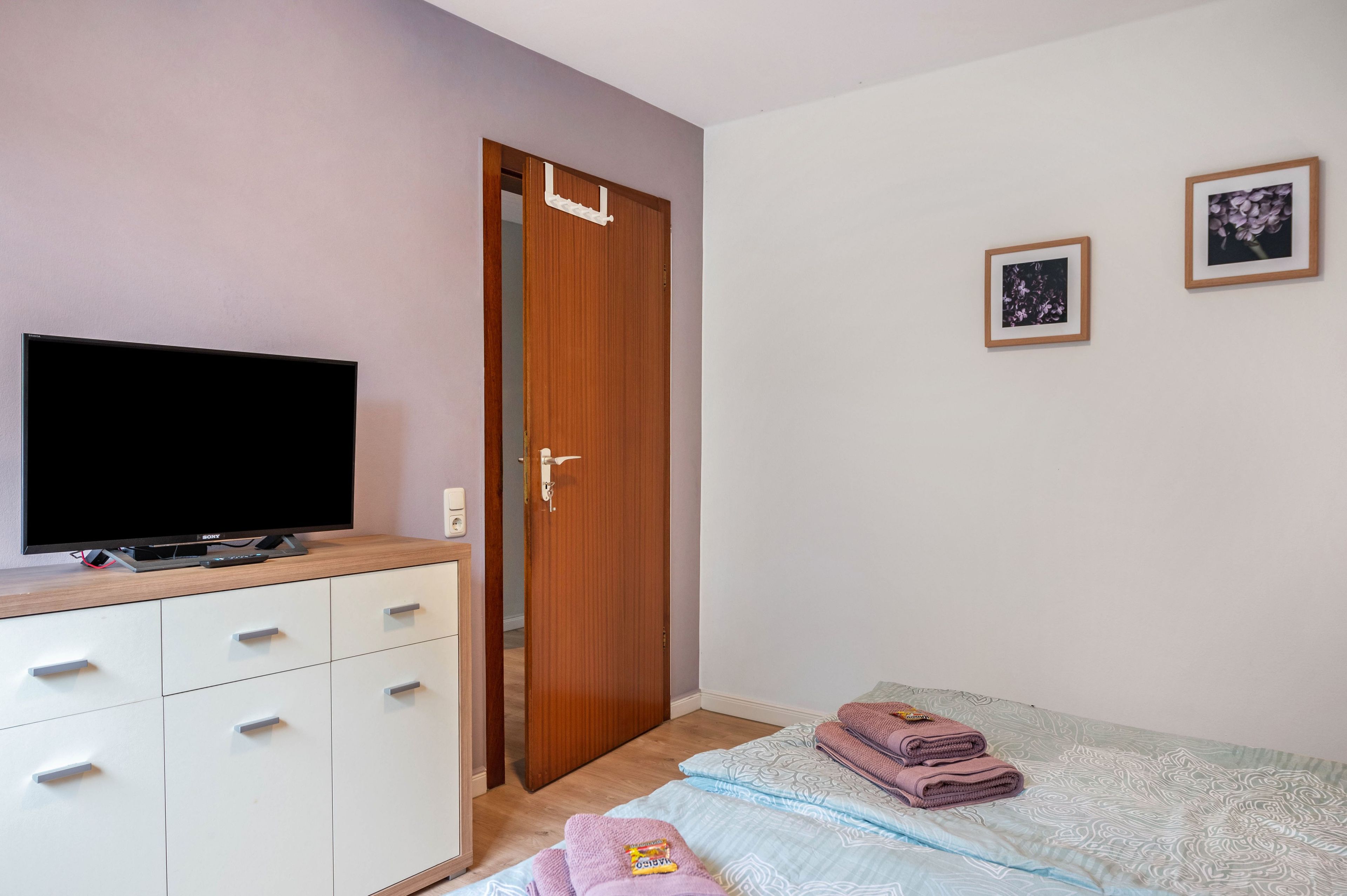 Ein Schlafzimmer mit einem Bett, einem Fernseher auf einer Kommode, einer Tür und zwei gerahmten Bildern an der Wand.