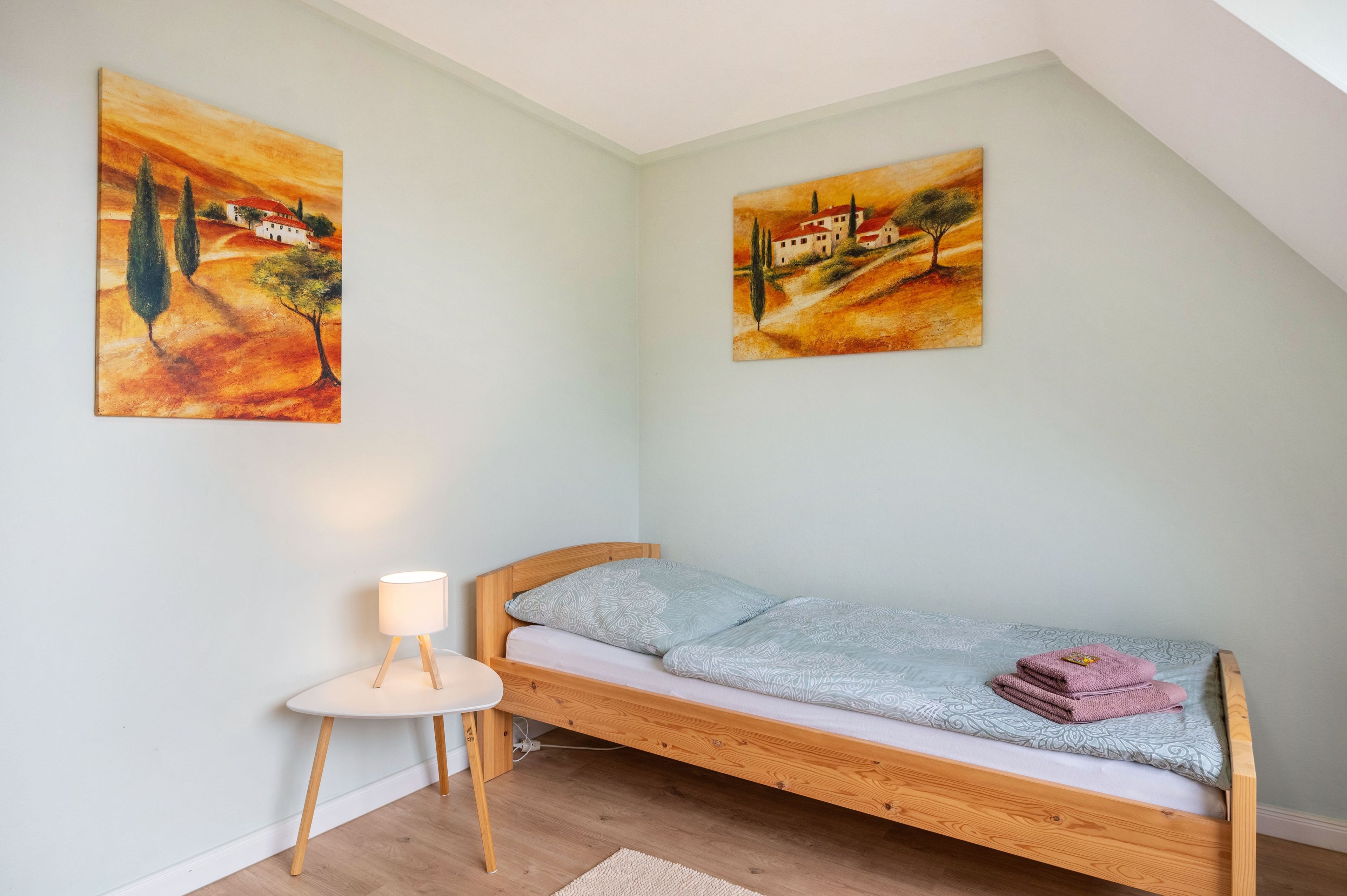 Uma cama de solteiro com estrutura de madeira e cabeceira branca, uma pequena mesa de cabeceira com abajur, e duas pinturas na parede.